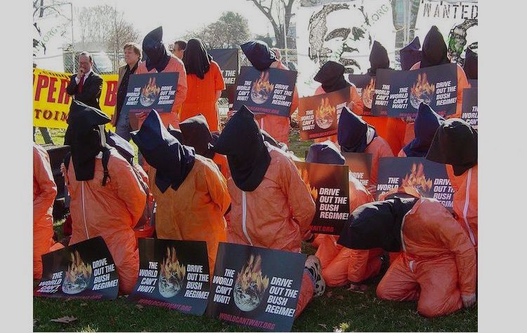Photo: Participants in a rally sponsored by The World Can't Wait are dressed as hooded detainees and holding WCW signs in Upper Senate Park on January 4, 2007. Photo by Ben Schumin. Source: Wikimedia Commons.