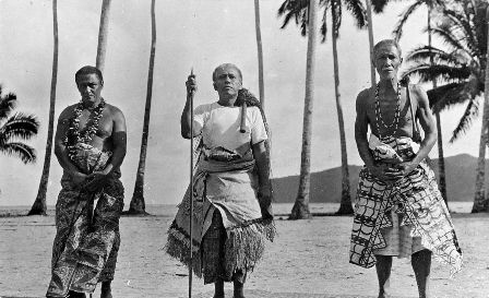 Three matai, the two older men bearing the symbols of orator chief status – the fue (flywhisk made of organic sennit rope with a wooden handle) over their left shoulder. The central elder holds the orator's wooden staff (to'oto'o) of office and wears an 'ie toga, fine matting. The other two men wear tapa cloth with patterned designs/ Public Domain