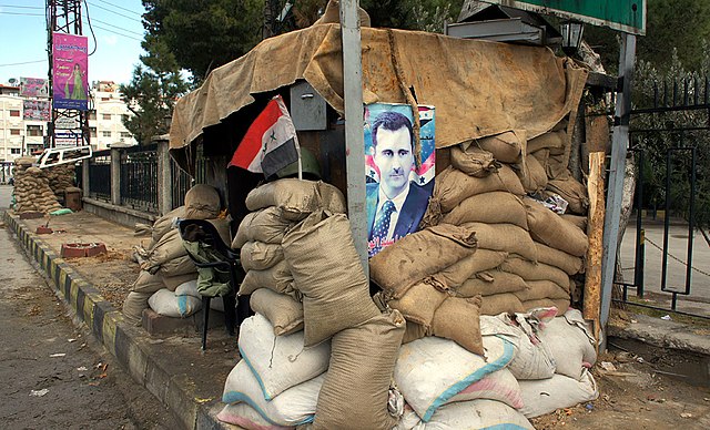 A poster of Syria's president at a checkpoint on the outskirts of Damascus, Jan. 14 2012./By Elizabeth Arrott - A View of Syria, Under Government Crackdown. VOA News photo gallery, Public Domain